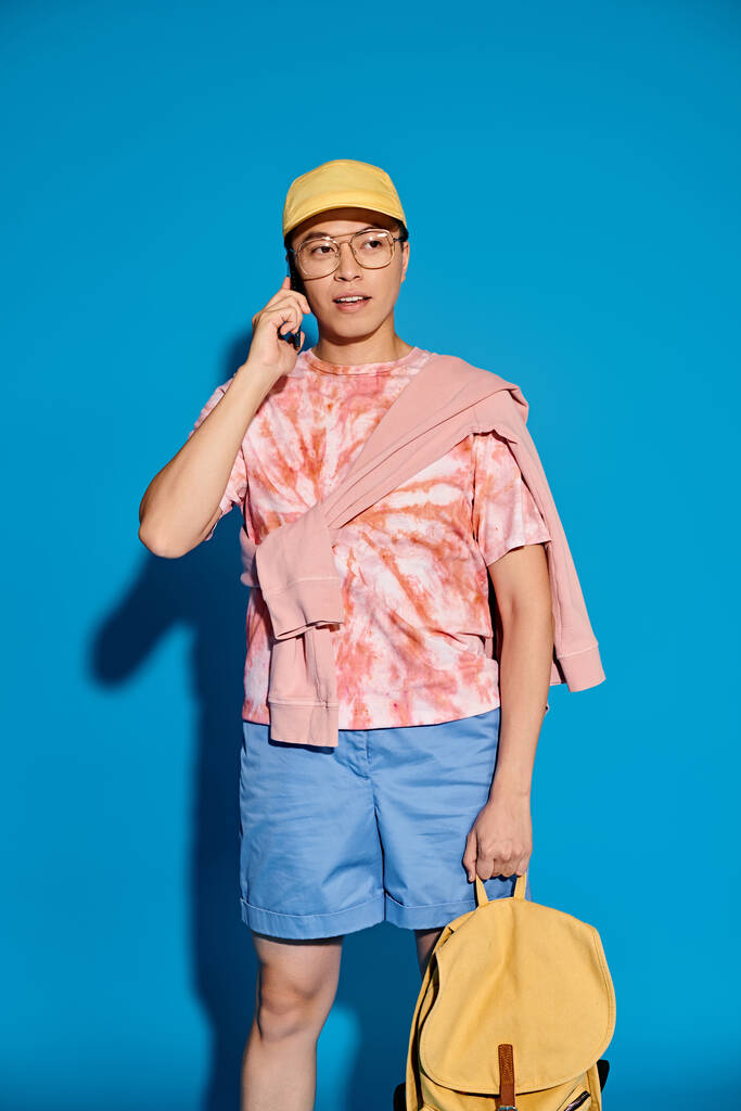 Un giovane elegante in abiti alla moda porta uno zaino e utilizza un telefono cellulare su uno sfondo blu. - Foto, immagini