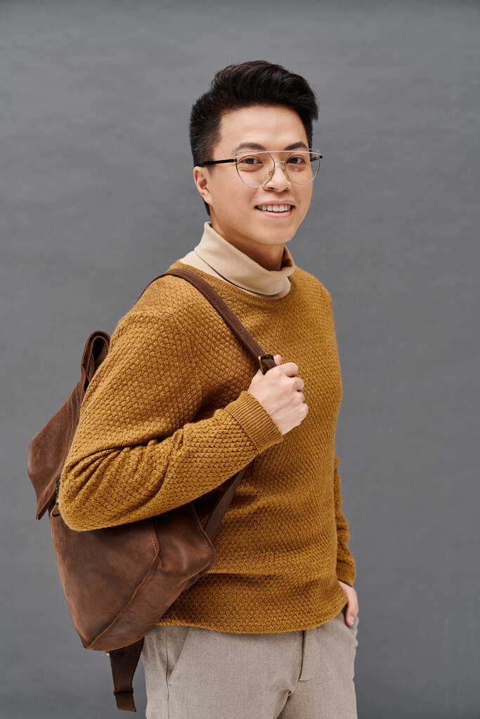 Egy divatos fiatalember szemüveggel és barna hátizsákkal magabiztosan pózol elegáns öltözékben.. - Fotó, kép