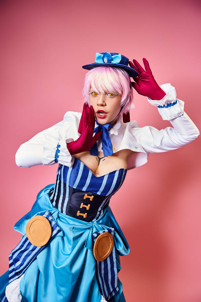 enticing sexy female cosplayer in blue hat and vibrant attire looking at camera on pink backdrop - Photo, Image