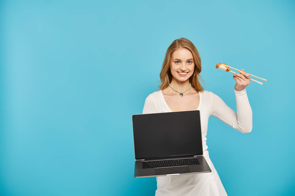 Een blonde vrouw met een laptop en Aziatisch eten, presentatie van een mix van technologie en culinaire hoogstandjes. - Foto, afbeelding