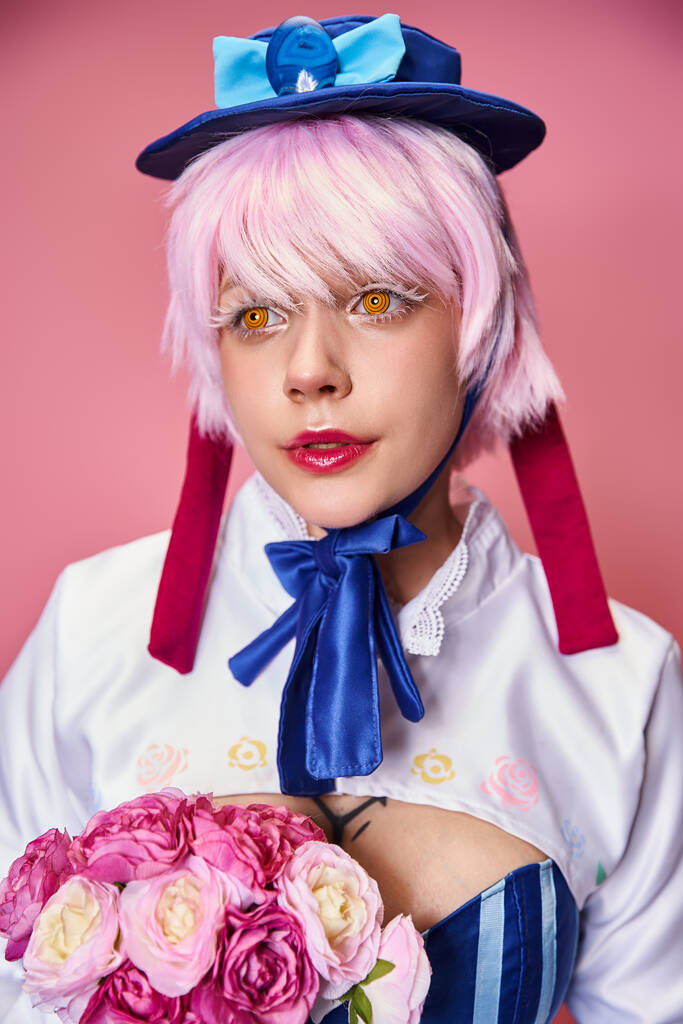 cosplayer femenino de moda en traje vívido sosteniendo flores rosadas y mirando hacia otro lado en el telón de fondo rosa - Foto, Imagen