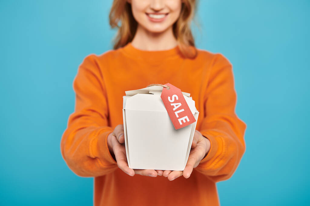 Vista recortada de mujer elegante sosteniendo caja de comida con una etiqueta de venta, mirando encantado e intrigado por el contenido. - Foto, imagen