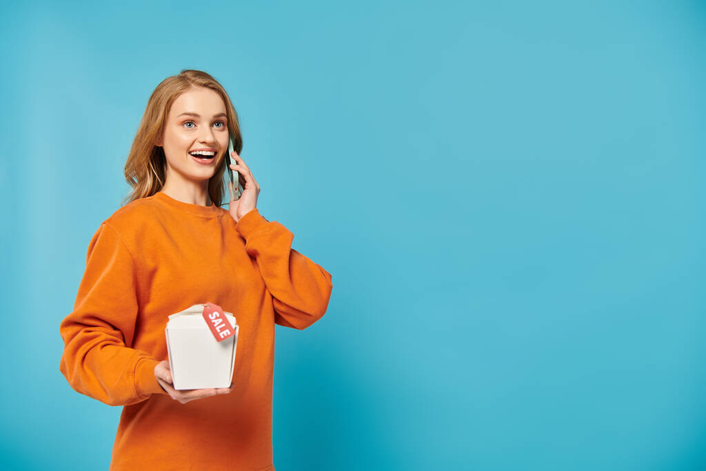 Eine stilvolle Frau im orangefarbenen Pullover führt ein lebhaftes Gespräch auf einem Handy mit einer Lebensmittelbox in der Hand. - Foto, Bild