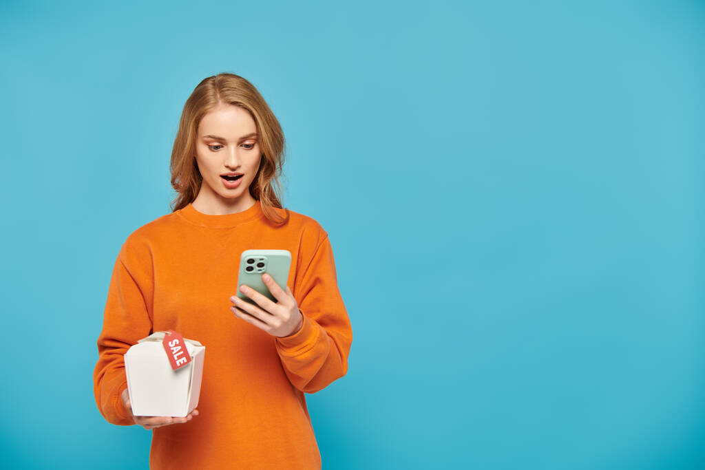 Une femme élégante dans un pull orange tient un téléphone portable en toute confiance et la nourriture boz étiquette de vente de dents sur elle. - Photo, image