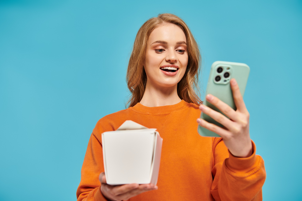 Een blonde vrouw poseert stilistisch balanceren een doos van Aziatische eten en een mobiele telefoon in haar handen. - Foto, afbeelding
