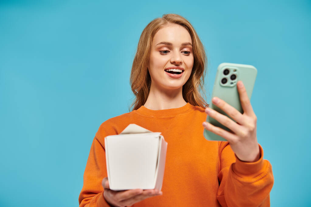 Une femme blonde pose élégamment en équilibrant une boîte de nourriture asiatique et un téléphone portable dans ses mains. - Photo, image