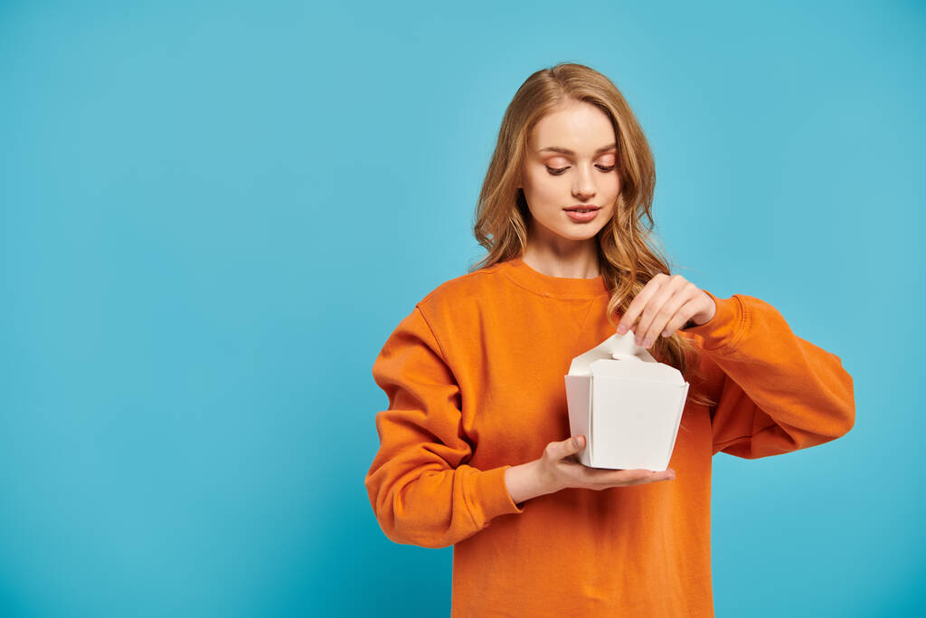 Eine schöne blonde Frau im orangefarbenen Pullover hält eine weiße Lebensmittelbox in der Hand. - Foto, Bild
