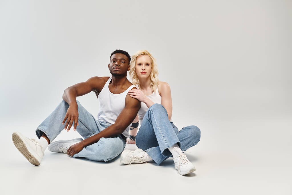 Una giovane coppia multiculturale seduta a terra in uno studio, che incarna una pacifica unione in mezzo a uno sfondo grigio. - Foto, immagini