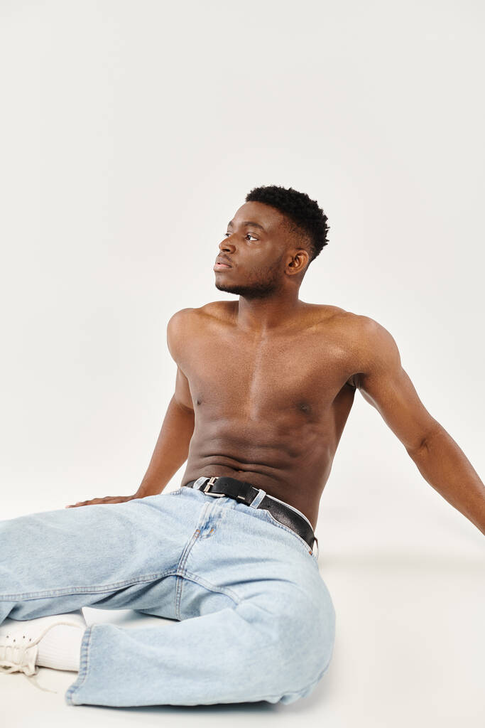 Muž bez trička Afroameričan ukazuje sílu a sebevědomí, když sedí na zemi ve studiu na šedém pozadí. - Fotografie, Obrázek