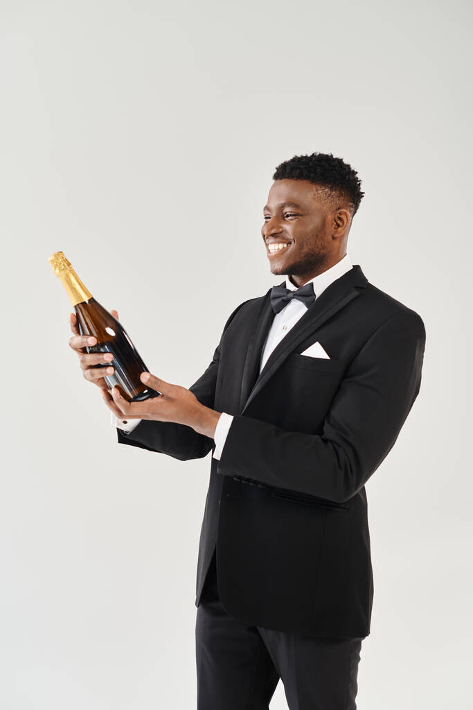 Handsome African American groom in a stylish tuxedo, holding a bottle of champagne, exuding elegance in a studio setting. - Photo, Image