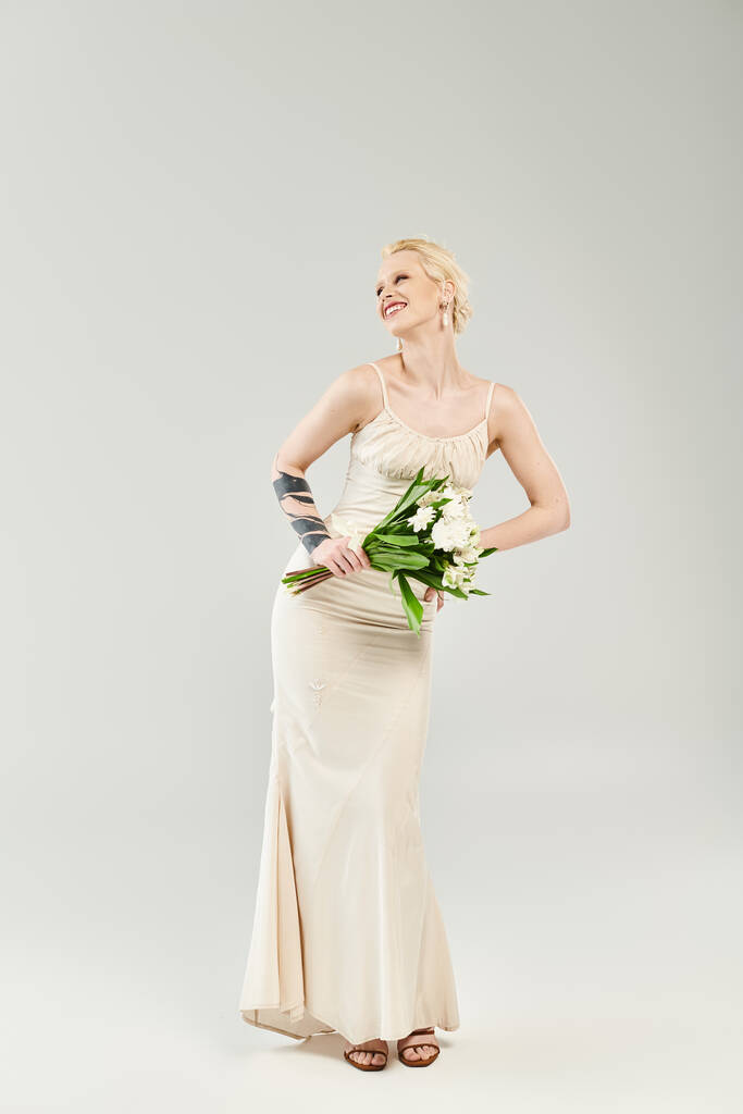 Een prachtige blonde bruid in een trouwjurk sierlijk houden van een levendige boeket bloemen tegen een grijze achtergrond. - Foto, afbeelding
