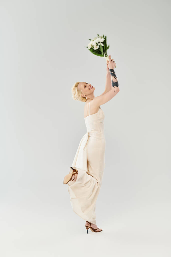 Eine schöne blonde Braut in einem weißen Kleid hält zart einen Blumenstrauß in einem Atelier vor grauem Hintergrund. - Foto, Bild