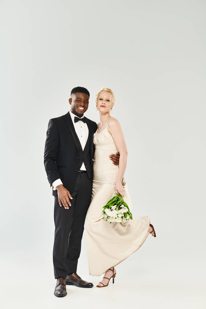 Een mooie blonde bruid in een trouwjurk en een Afro-Amerikaanse bruidegom, beide in formele kleding, elegant poserend voor een portret in een studio op een grijze achtergrond. - Foto, afbeelding