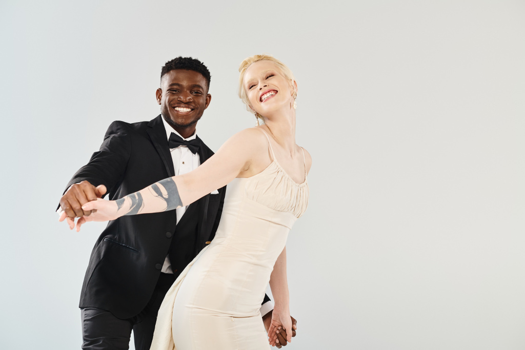 Красивая блондинка невеста в свадебном платье стоит рядом с афроамериканским женихом в остром смокинге на сером фоне. - Фото, изображение