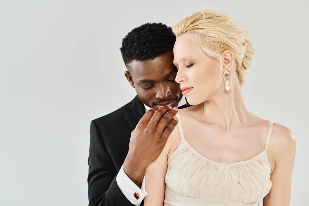 Une belle mariée blonde dans une robe de mariée se tient à côté de son marié afro-américain dans un studio sur un fond gris. - Photo, image