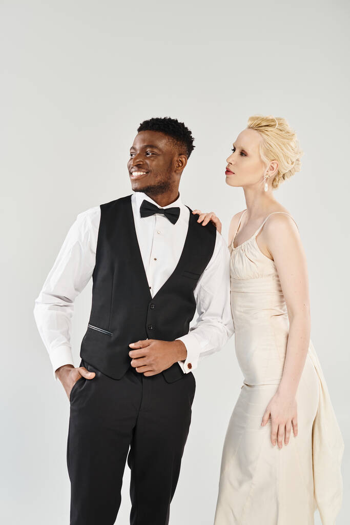 Una hermosa novia rubia en un vestido de novia blanco está junto a su novio afroamericano en un esmoquin, exudando elegancia y amor. - Foto, imagen