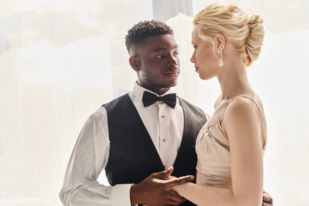 Piękna blond panna młoda w sukni ślubnej i afro-amerykański pan młody stojący razem w studio na szarym tle. - Zdjęcie, obraz