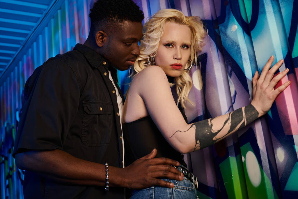 Eine blonde Frau und ein afroamerikanischer Mann stehen Seite an Seite auf einer städtischen Straße mit bunten Graffiti an den Wänden. - Foto, Bild