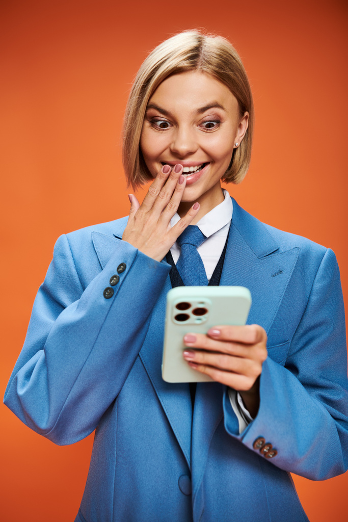 θετική καλοντυμένη γυναίκα με κοντά ξανθά μαλλιά σε σικ ενδυμασία κρατώντας τηλέφωνο σε πορτοκαλί φόντο - Φωτογραφία, εικόνα