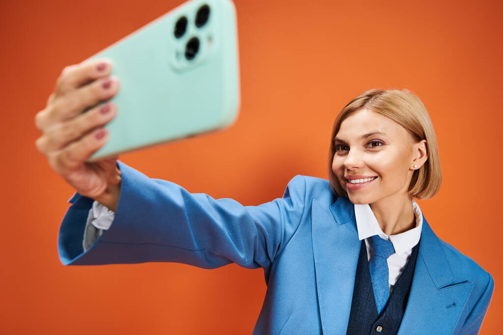 щаслива добре одягнена жінка з коротким світлим волоссям у шикарному вбранні, що тримає телефон на помаранчевому тлі - Фото, зображення