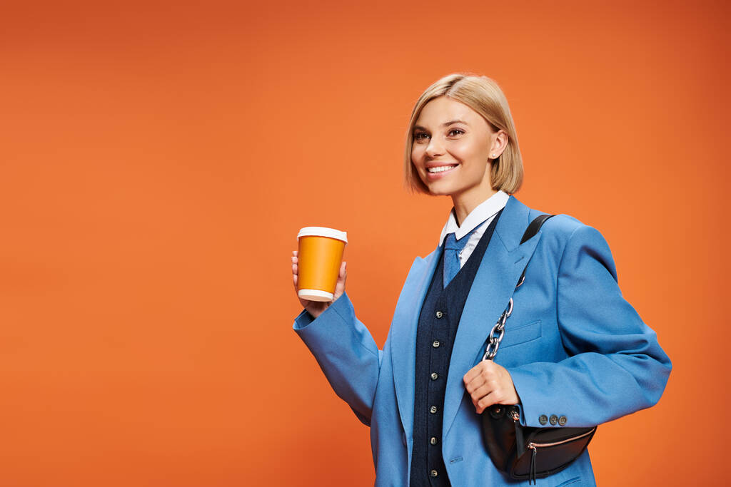 mujer sofisticada alegre con pelo corto rubio sosteniendo bolso y café sobre fondo naranja - Foto, imagen
