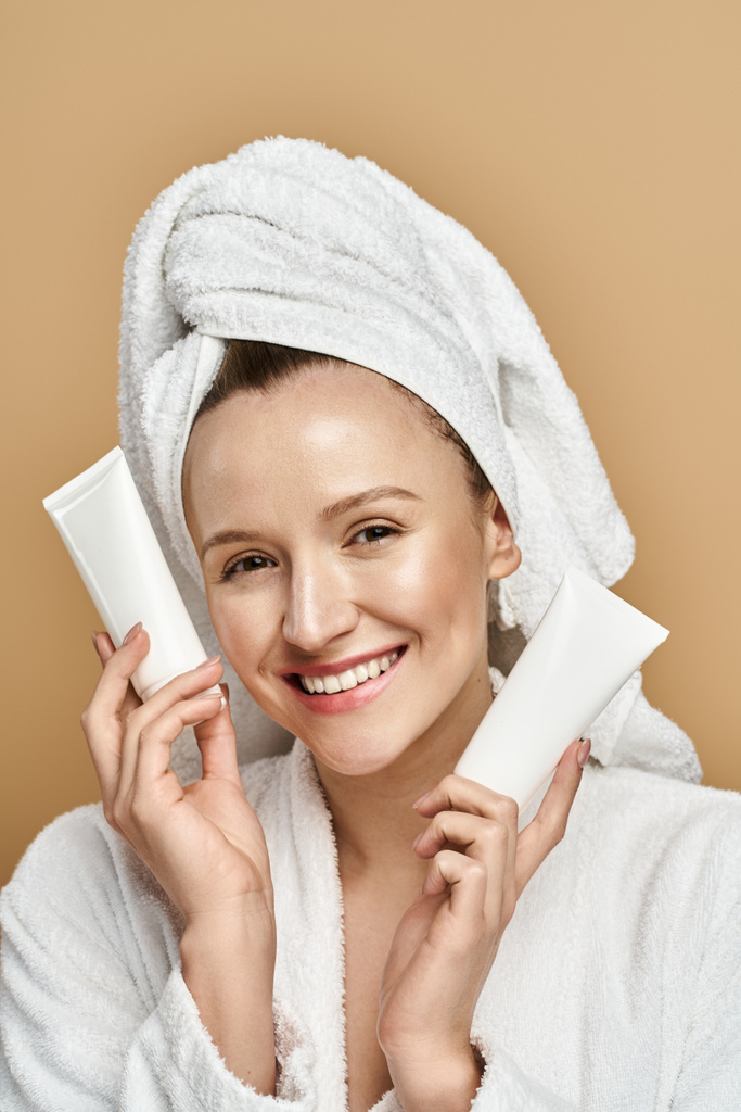 Une femme avec une serviette enroulée autour de sa tête tient un tube de crème, mettant en valeur sa routine beauté. - Photo, image