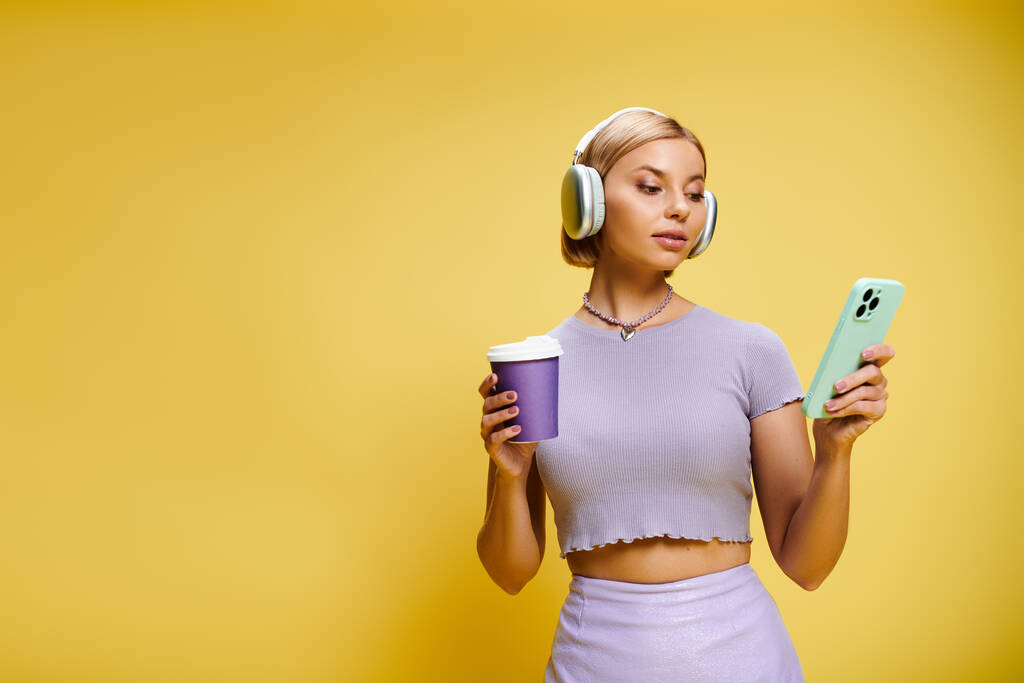 atraente mulher alegre com fones de ouvido e telefone celular desfrutando de seu café no fundo amarelo - Foto, Imagem