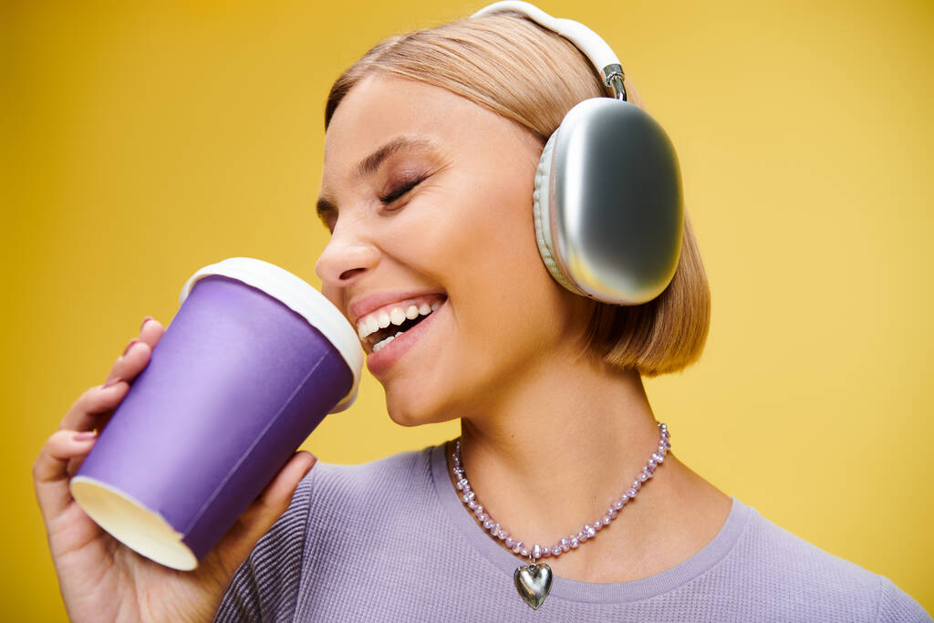 joyous stylish woman with headphones enjoying music and hot coffee while posing on yellow backdrop - Photo, Image