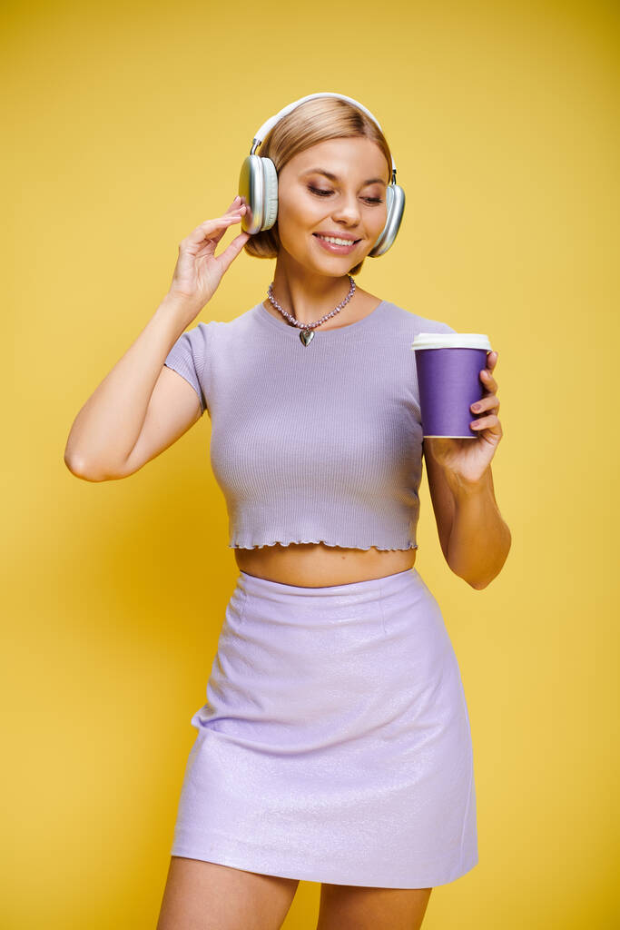 vidám elegáns nő fejhallgatóval élvezi a zenét és a forró kávét, miközben pózol a sárga háttérben - Fotó, kép