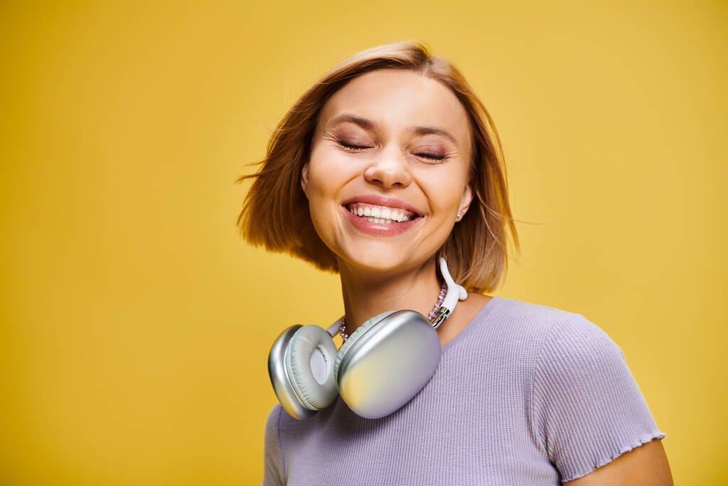 黄色い背景で音楽を楽しんでいる短いブロンドの髪とヘッドフォンを持つかなり楽しい女性 - 写真・画像