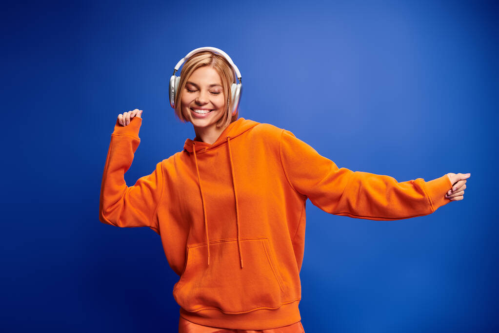 Kısa sarı saçlı ve kulaklıklı çekici bir kadın mavi arka planda müzik dinliyor. - Fotoğraf, Görsel