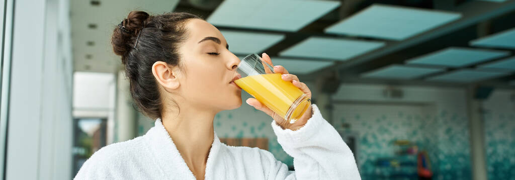 Młoda, piękna brunetka delektująca się szklanką orzeźwiającego soku pomarańczowego w krytym spa z basenem. - Zdjęcie, obraz