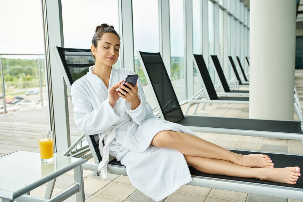 Een jonge brunette vrouw in een badjas ontspant in een indoor spa terwijl met behulp van een mobiele telefoon. - Foto, afbeelding