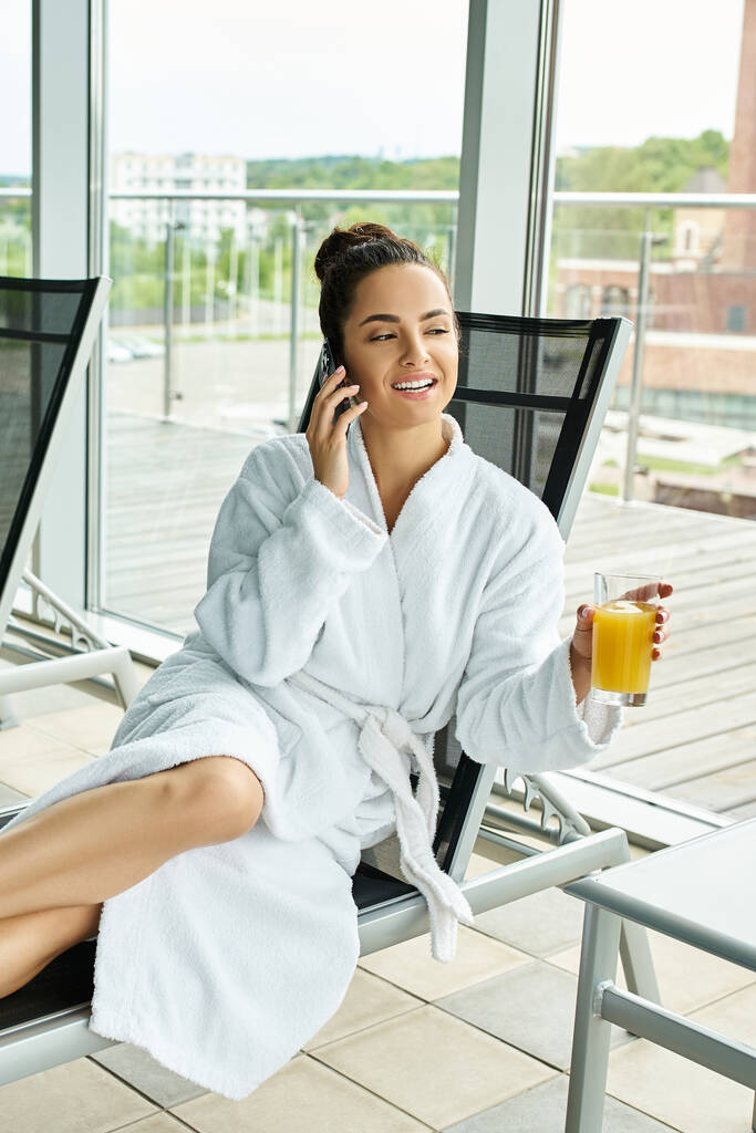 A young brunette woman is sitting in a lounge chair by an indoor spa pool, engaged in a phone conversation. - Photo, Image