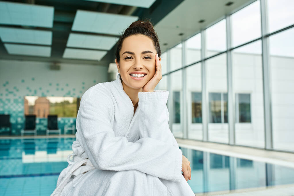 Uma jovem morena de roupão de banho desfrutando de um momento relaxante junto à piscina em um spa interior. - Foto, Imagem