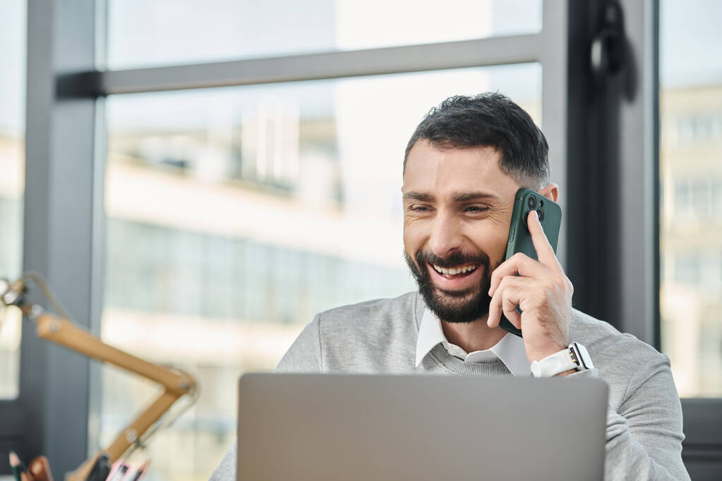 Ein Mann sitzt vor einem Laptop und telefoniert mit einem Handy, vertieft in seine Arbeit in einem geschäftigen Büro. - Foto, Bild