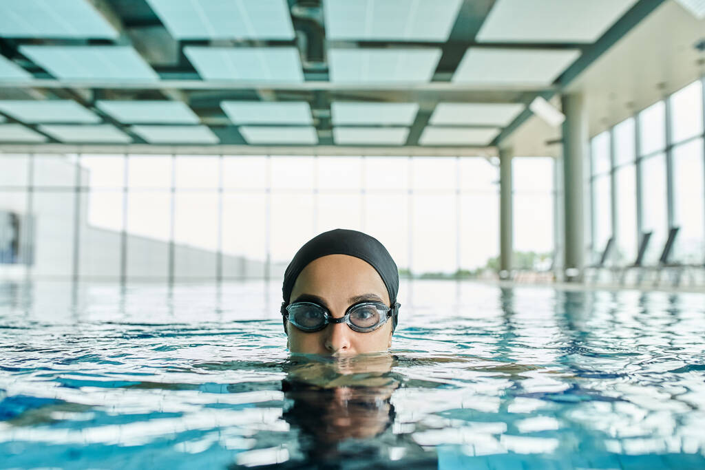 Mulher jovem desliza através da piscina em óculos e touca de natação, incorporando movimento fluido e tranquilidade. - Foto, Imagem