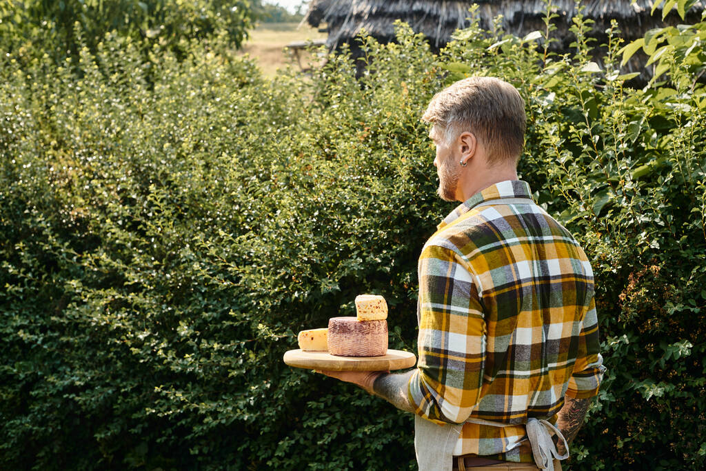 εμφανίσιμος άντρας με τατουάζ στα χέρια με casual ενδυμασία κρατώντας τυρί και κοιτάζοντας μακριά, ενώ στη φάρμα - Φωτογραφία, εικόνα