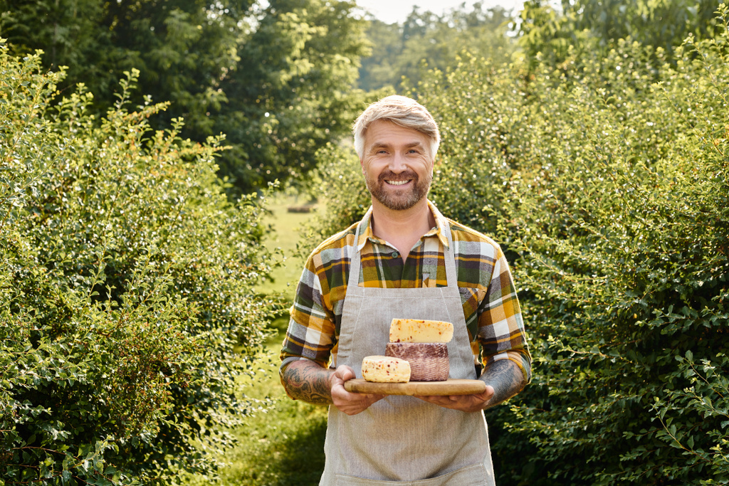 vreugdevolle knappe boer met tatoeages met zelfgemaakte kaas in zijn handen en glimlachend naar de camera - Foto, afbeelding