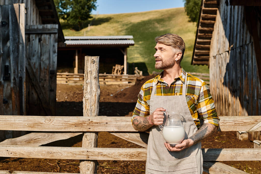 attractive man in everyday outfit with tattoos on arms holding jar of fresh milk and looking away - Photo, Image