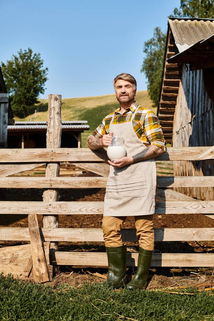 dobře vypadající muž s tetováním na pažích drží sklenici čerstvého mléka a při pohledu do kamery, zatímco na farmě - Fotografie, Obrázek