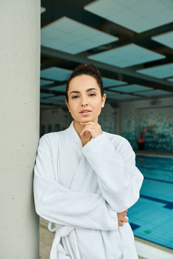 Een jonge vrouw in een badjas geniet van een rustig moment bij een overdekt zwembad in een spa-omgeving. - Foto, afbeelding