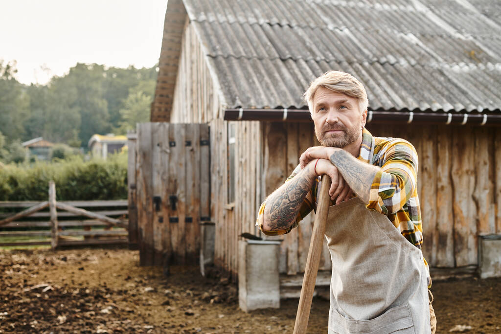 trabajador atractivo granjero con barba y tatuajes usando horquilla mientras se trabaja con estiércol - Foto, imagen