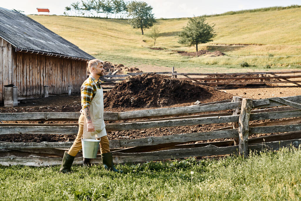 gut aussehender bärtiger Mann mit Tätowierungen auf den Armen, der Eimer mit frischer Milch hält, während er auf seinem Bauernhof ist - Foto, Bild