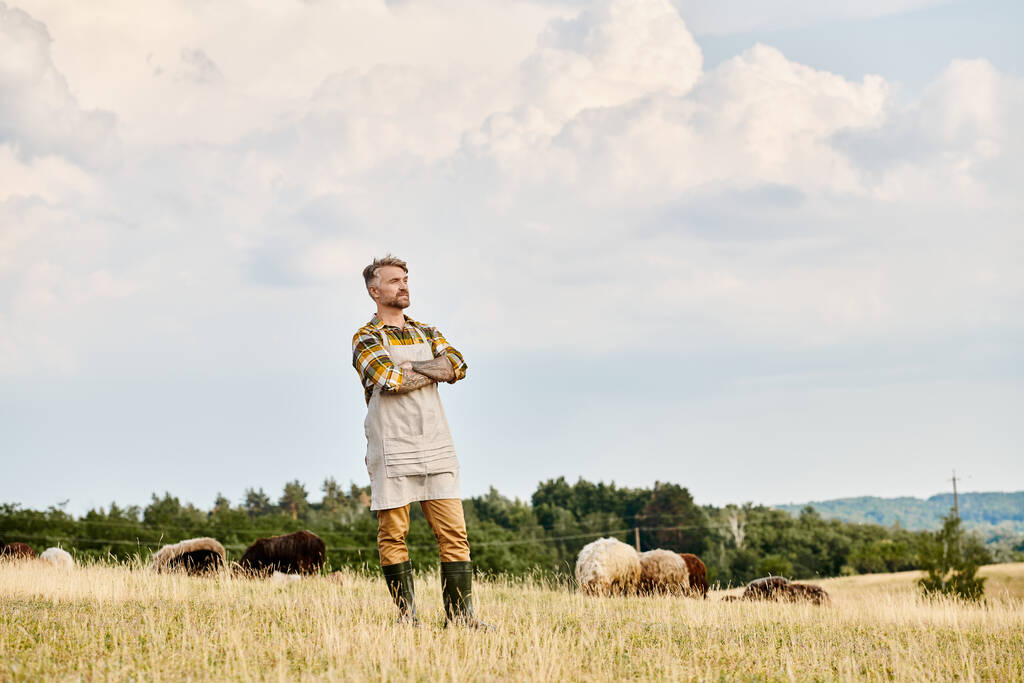 moderner bärtiger Bauer mit Tätowierungen, der mit verschränkten Armen posiert und mit Rindern im Hintergrund wegsieht - Foto, Bild