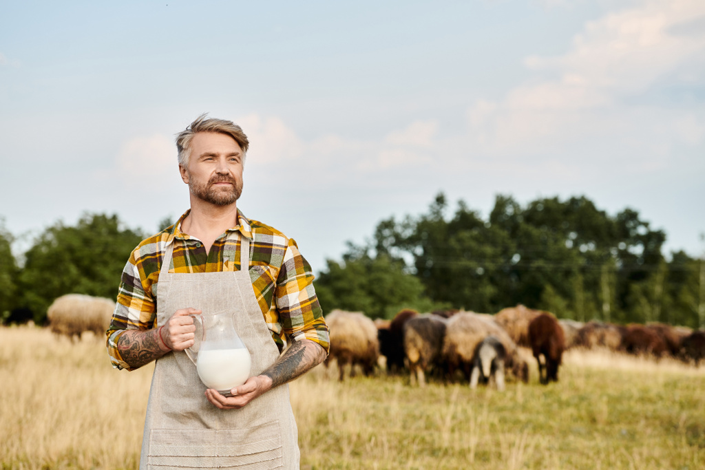 Dövmeleri olan yakışıklı, sakallı, modern bir çiftçi. Elinde arka planda koyunları olan taze süt kavanozu var. - Fotoğraf, Görsel