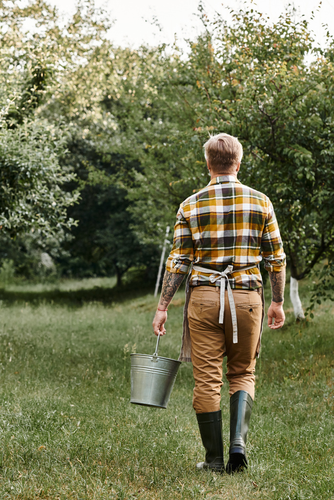 vertical shot of hard working man with tattoos working in garden and holding metal bucket in hand - Photo, Image