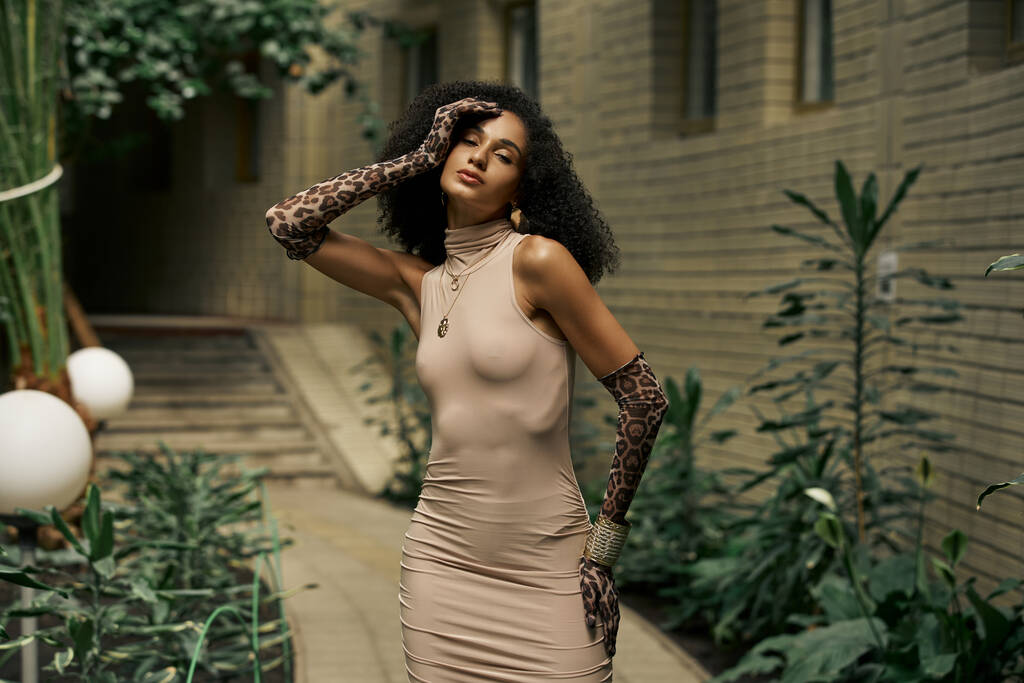 Geavanceerde en krullende zwarte vrouw in jurk en dierlijke print handschoenen poseren in stedelijke tuin - Foto, afbeelding