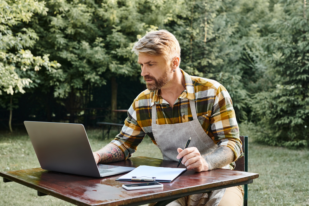 hart arbeitender gutaussehender Mann mit Bart mit Laptop und Klemmbrett, um Ressourcen auf dem Bauernhof zu analysieren - Foto, Bild