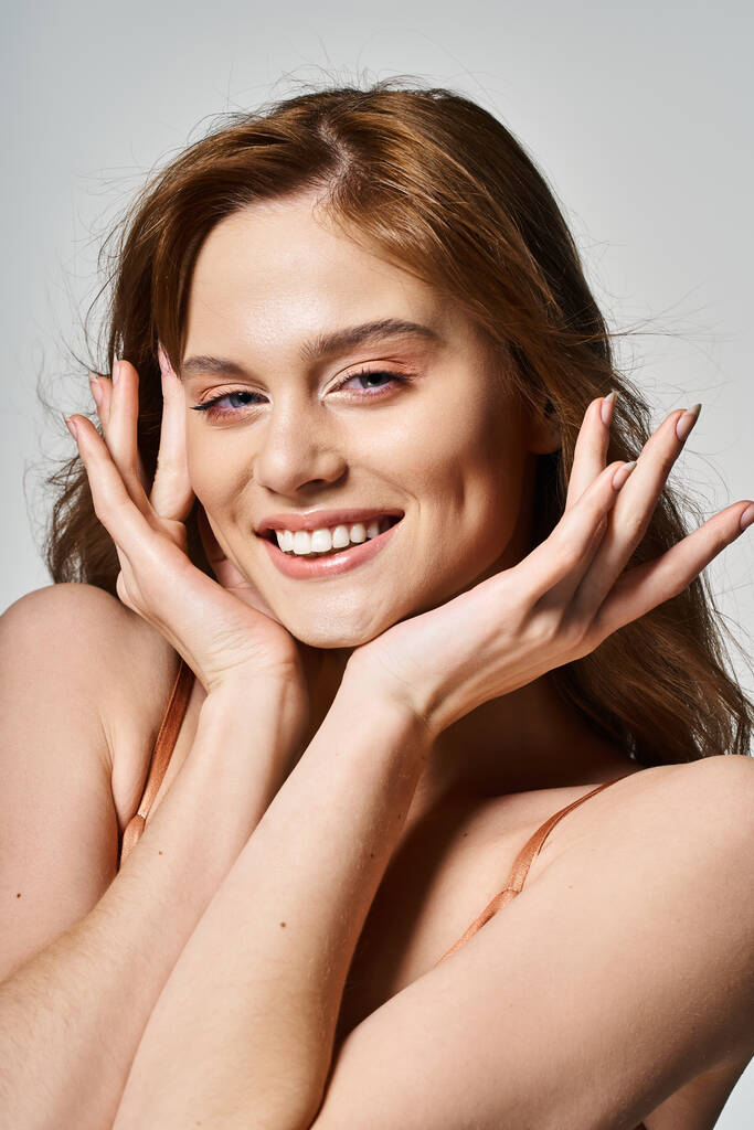 Vrolijke glimlachende vrouw met trendy make-up, kijkend naar de camera, handen in de buurt van gezicht op grijze achtergrond - Foto, afbeelding